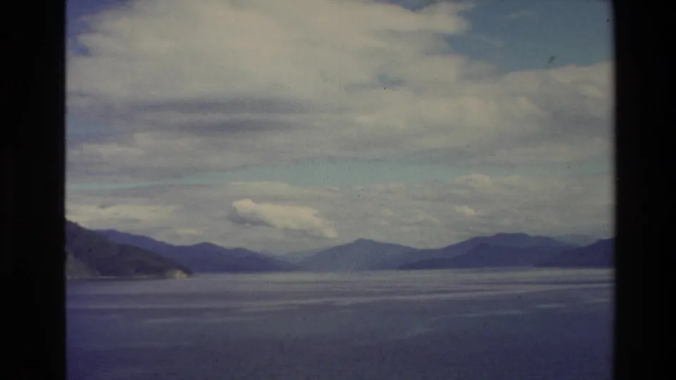 1982 New Zealand 8mm frame with open blue water, layered distant mountains, low clouds, and visible dark film edges from the scan.