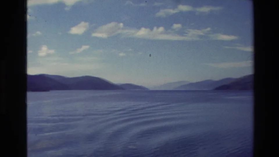 New Zealand coastal water stretches toward distant hills beneath a bright sky in 1982 8mm home-movie footage.