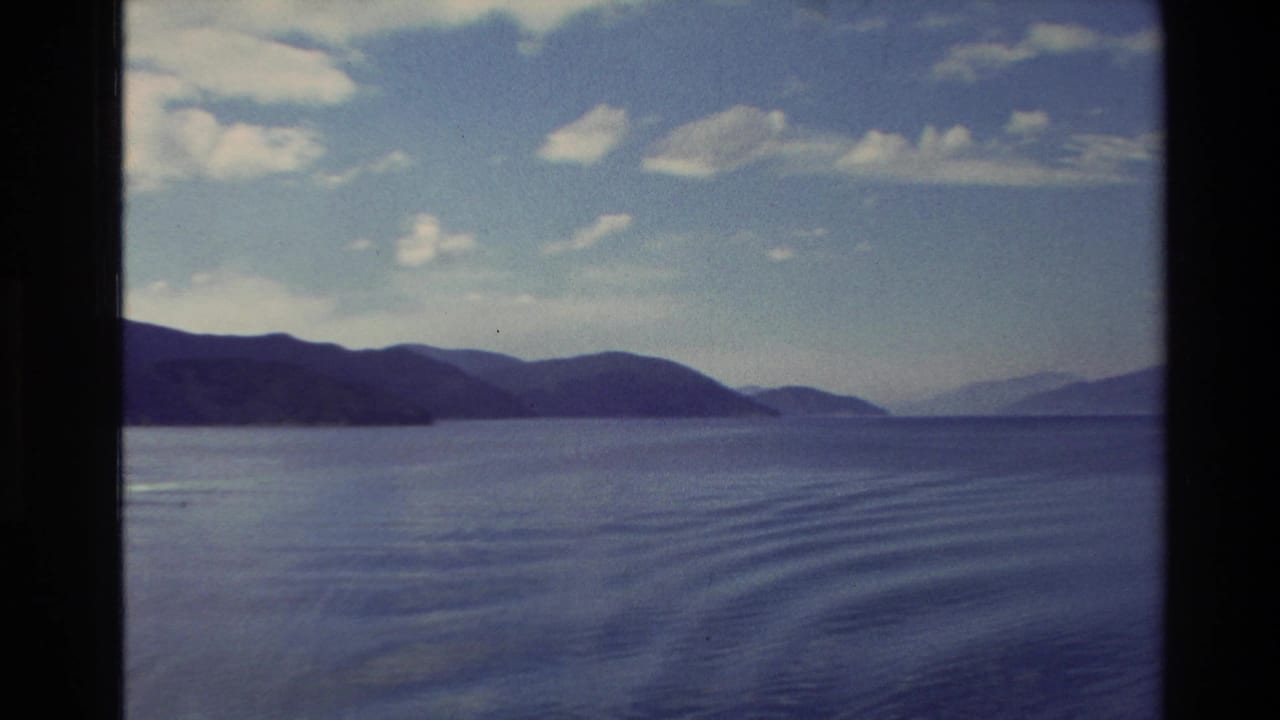 1982 New Zealand 8mm frame showing calm lake water, distant mountain ridges, and a partly cloudy sky from a moving vessel.