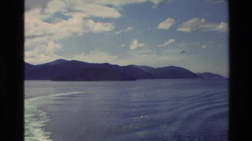 1982 New Zealand 8mm frame with a mountain ridge, open lake water, clouded sky, and wake patterns trailing across the surface.