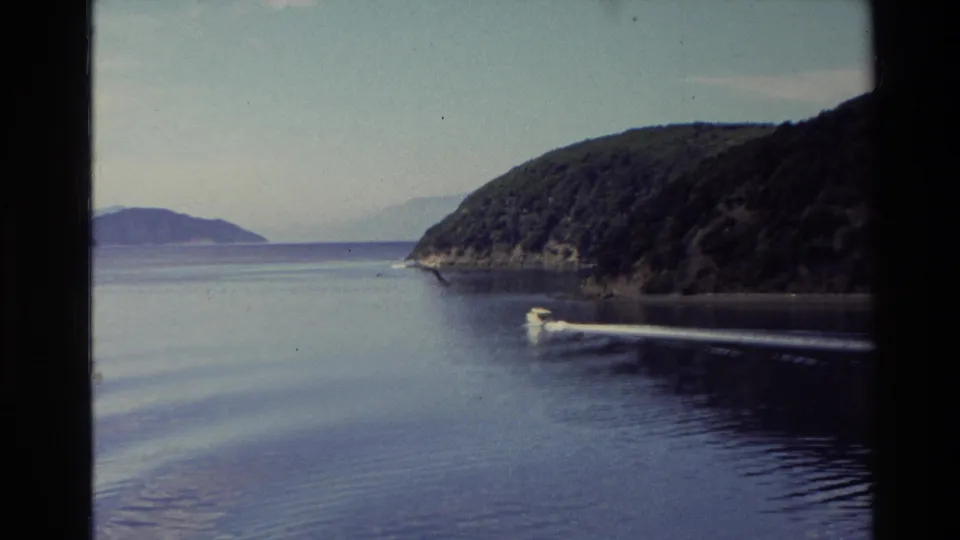 1982 New Zealand 8mm frame showing a small motorboat skimming past wooded shoreline on calm lake water with distant hills beyond.