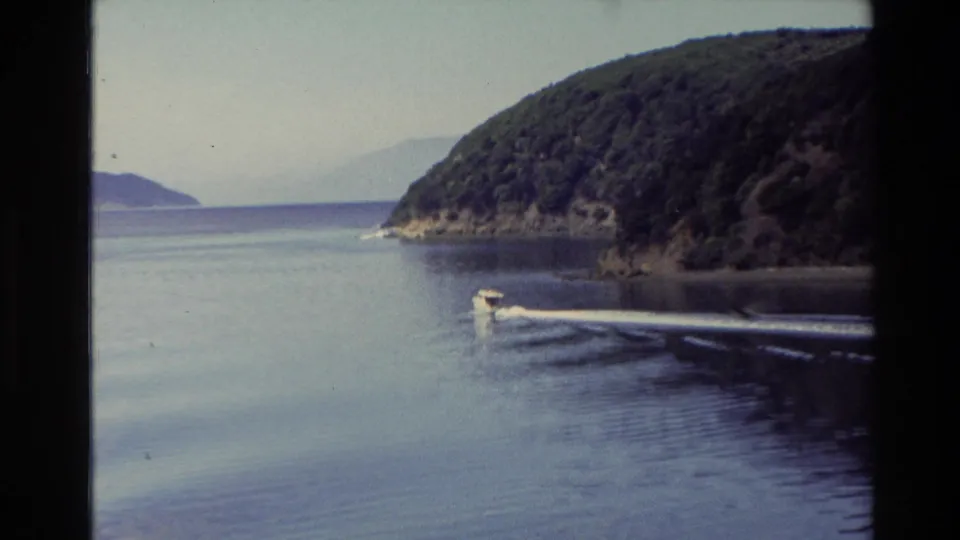 1982 New Zealand 8mm frame showing a motorboat and white wake beside a wooded headland, with calm water and hazy hills in the distance.
