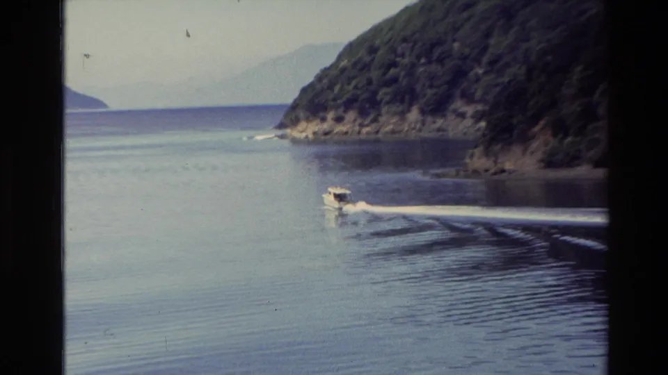 1982 New Zealand 8mm frame showing a small motorboat skimming across calm lake water below wooded hills.