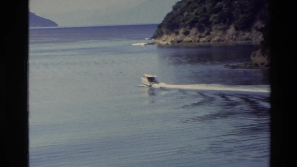 1982 New Zealand 8mm frame with a small motorboat crossing open lake water between wooded shorelines and distant hills.
