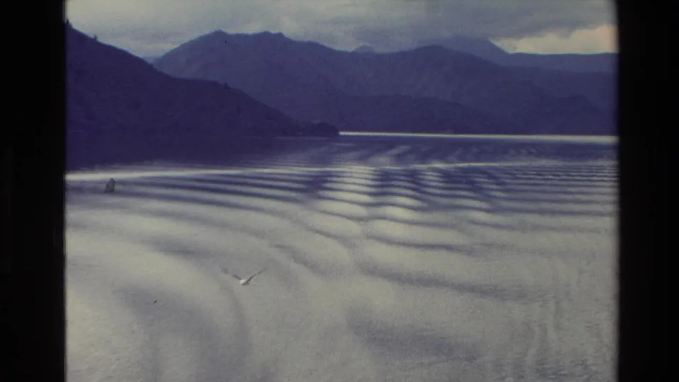 Wide 1982 New Zealand lake view with layered mountains, wake ripples across the water, and a small bird near the surface.