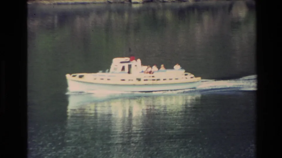 A white motorboat with distant passengers moves across New Zealand lake water in a soft 1982 8mm home-movie frame.