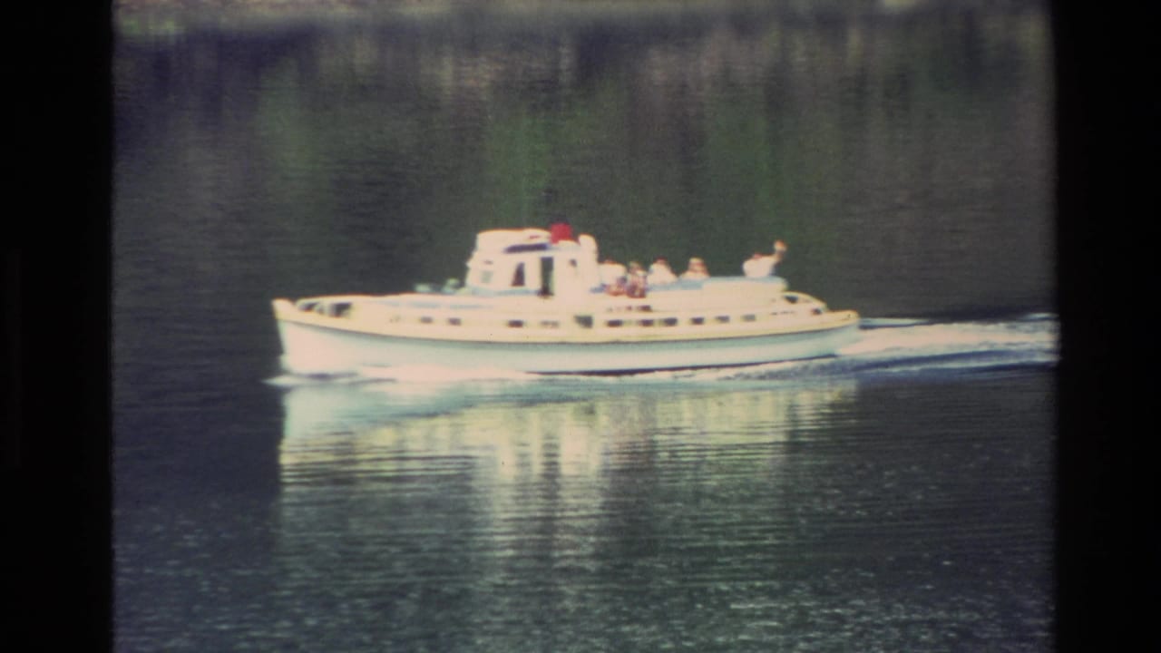 A white motorboat moves across calm New Zealand water in 1982 home-movie footage, with several passengers visible on deck.