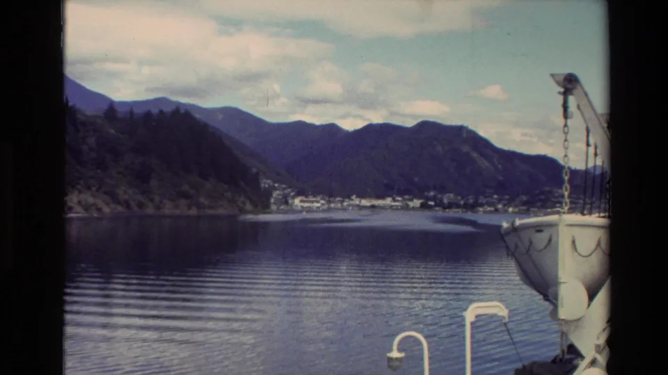 Mountain-backed shoreline and a small New Zealand town appear across calm water, with part of a vessel visible at the right edge.