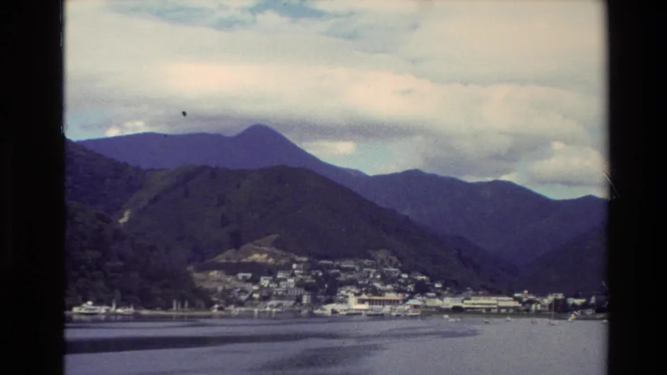 A small New Zealand waterfront town is visible beneath rounded green hills and layered mountains in faded 8mm color.