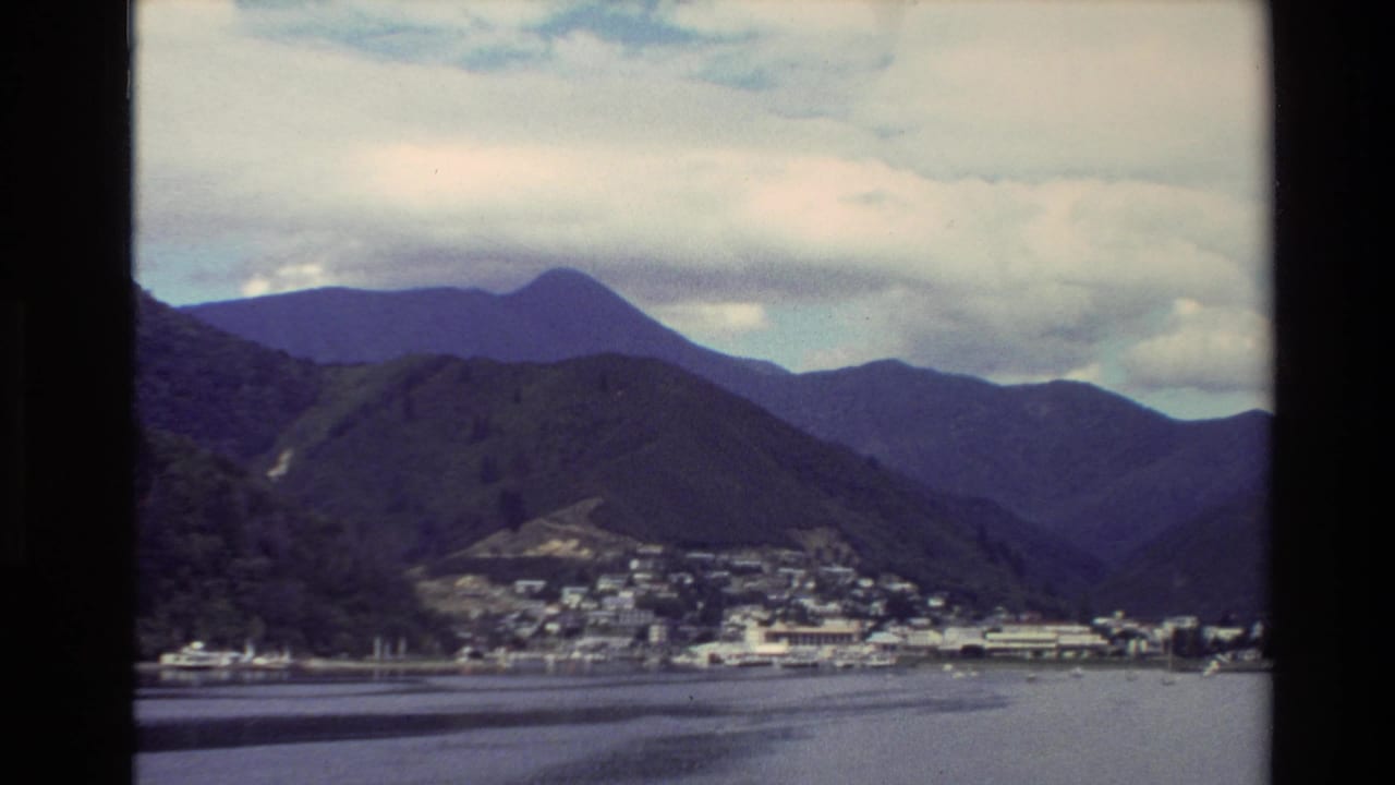 1982 New Zealand home-movie view of a coastal town at the base of green mountains beside still harbor water.