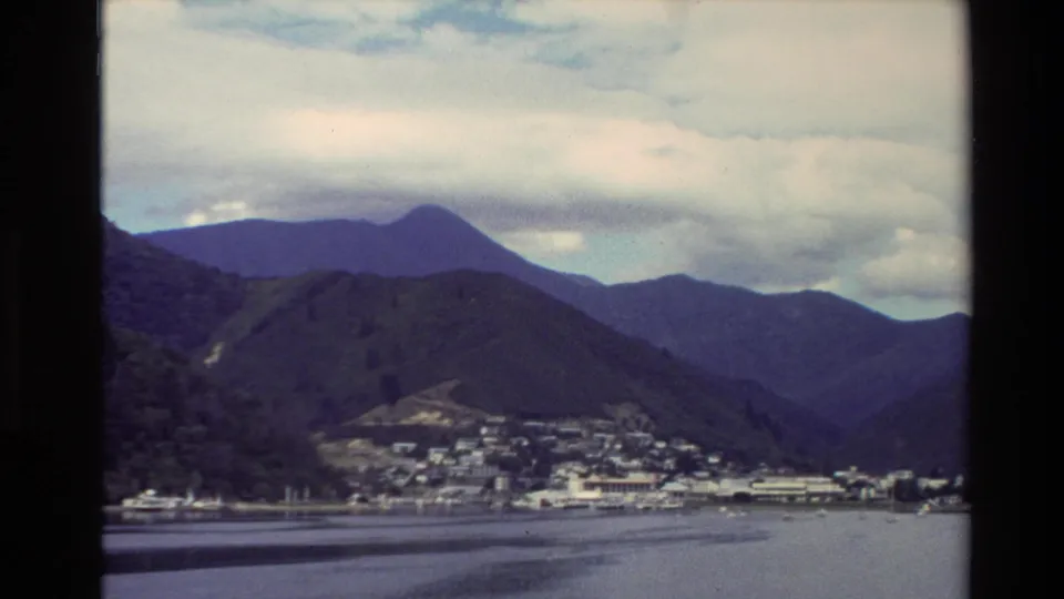 1982 New Zealand home-movie view of a coastal town at the base of green mountains beside still harbor water.