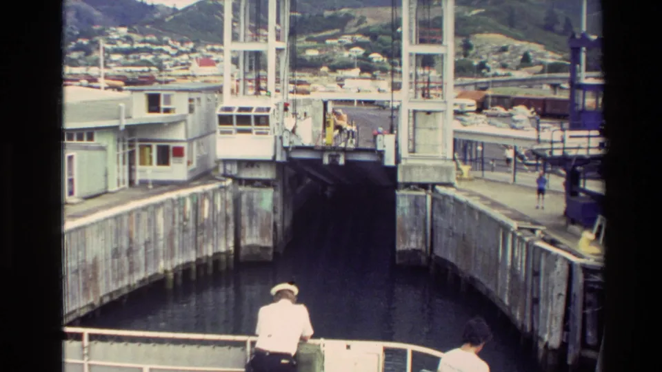 Ferry or vessel deck view approaching a bridge-like harbor structure in New Zealand, with distant people and waterfront buildings visible.