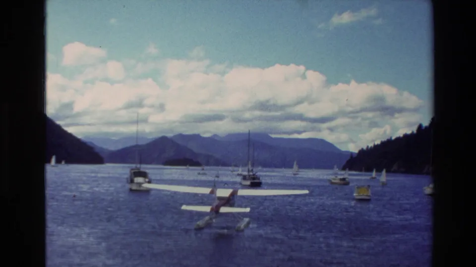 1982 New Zealand harbor or lake scene with a seaplane floating among sailboats beneath bright clouds and distant mountains.