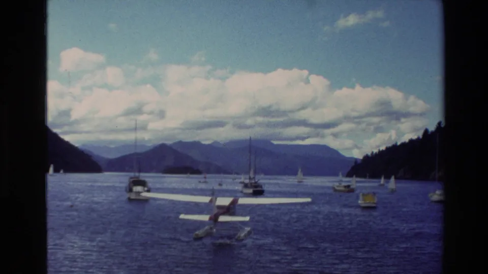 New Zealand 1982 home-movie frame of a seaplane on open water near moored sailboats, with layered mountains under a blue sky.