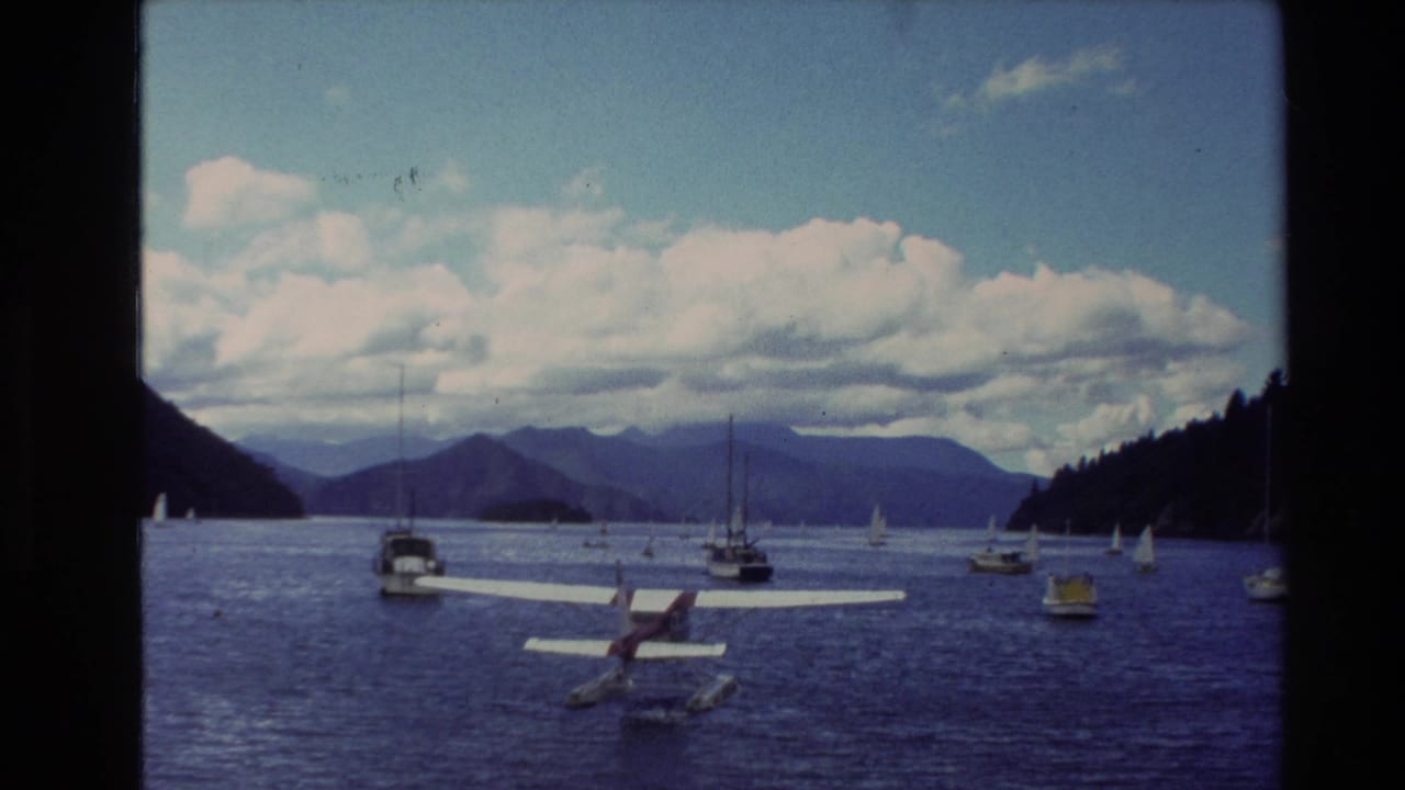 1982 New Zealand 8mm frame showing a seaplane on a lake, scattered sailboats, wooded slopes, distant mountains, and cloudy sky.