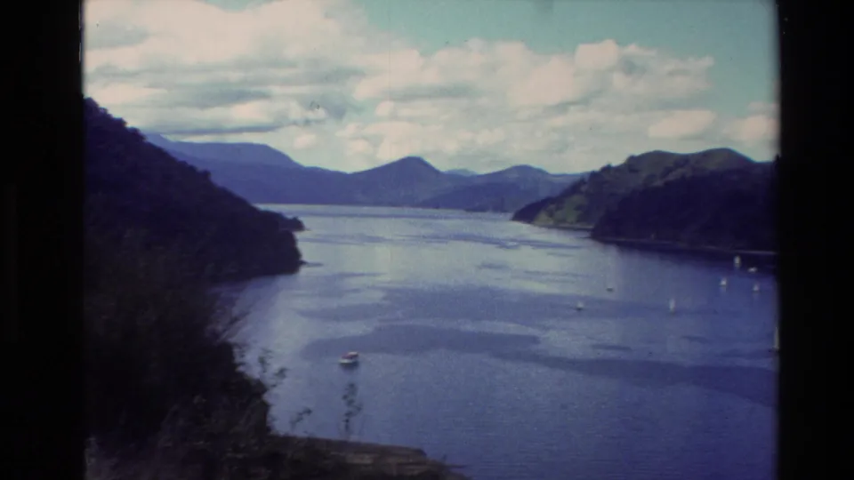 1982 New Zealand home-movie frame looking across a broad lake toward wooded hills, distant mountains, small boats, and a partly cloudy sky.