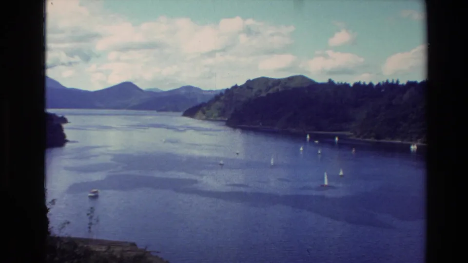 1982 New Zealand 8mm frame showing sailboats spread across a calm lake channel, with wooded shoreline and layered mountains beyond.