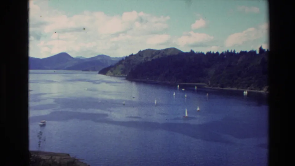 1982 New Zealand home-movie frame of a forest-lined lake with several sailboats, a small boat, distant mountains, and visible 8mm film border.
