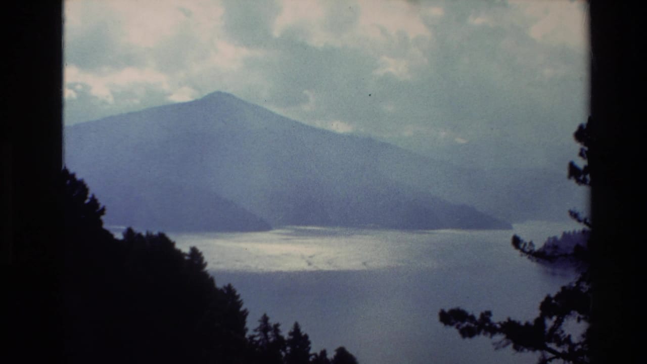 1982 New Zealand home-movie frame showing hazy mountains across calm water, with dark trees in the foreground.