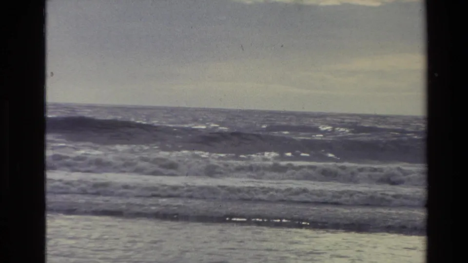 1982 New Zealand coastal frame with low rolling surf, a flat horizon, and muted cloudy light over the sea.