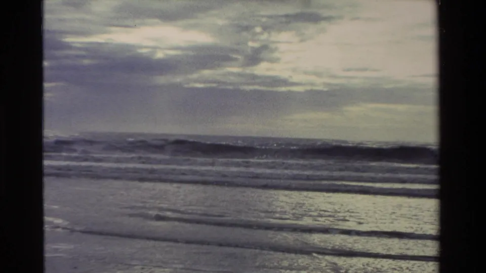 1982 New Zealand home-movie frame showing wet sand, layered surf lines, and a bright overcast sky at the coast.