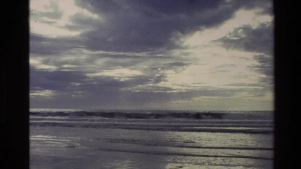 1982 New Zealand coastal frame with dramatic cloud cover, distant surf, and reflective wet shoreline in soft 8mm color.