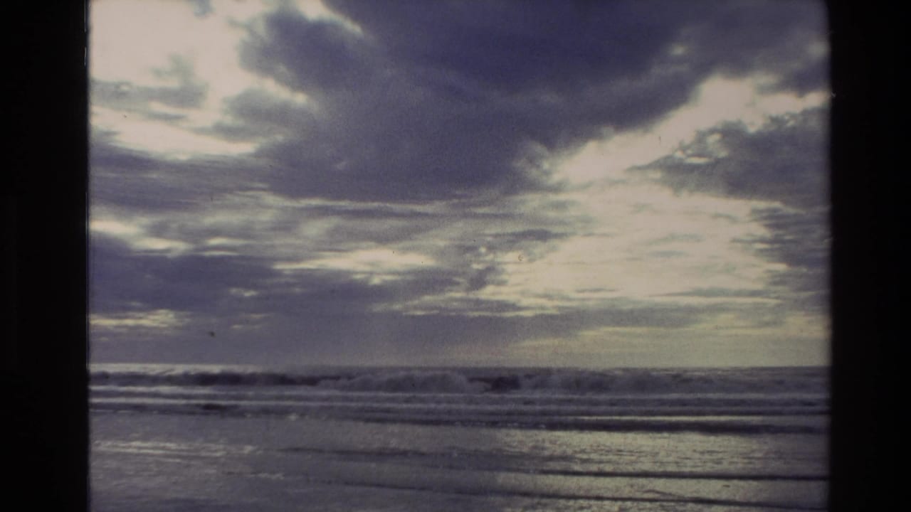 1982 New Zealand 8mm frame showing a cloudy coastal horizon, low surf, and sun filtering through thick cloud over the water.