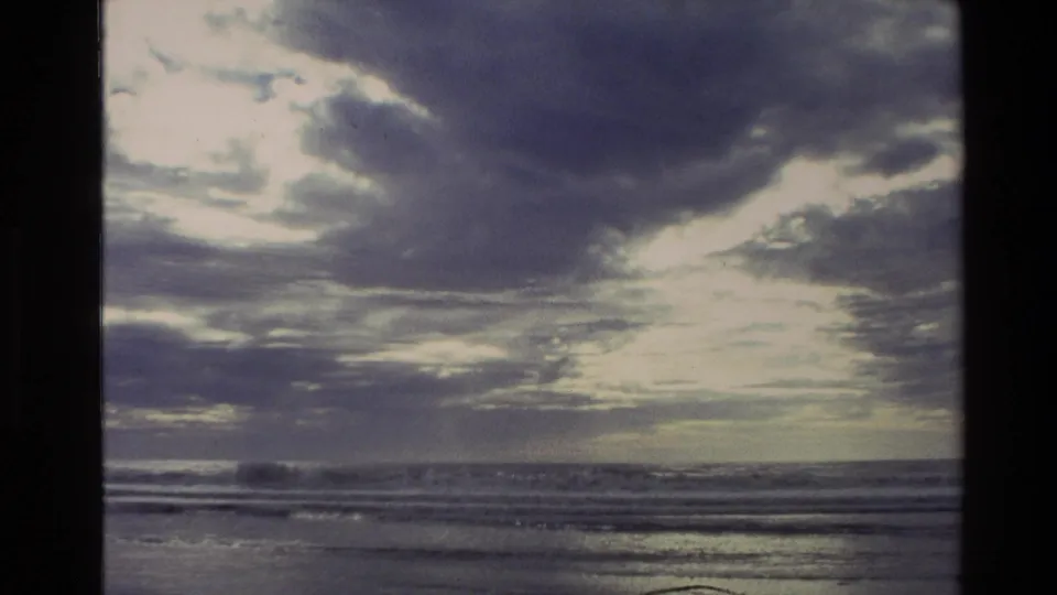 1982 New Zealand 8mm frame with bright cloud breaks over a dark ocean horizon and small waves moving toward shore.