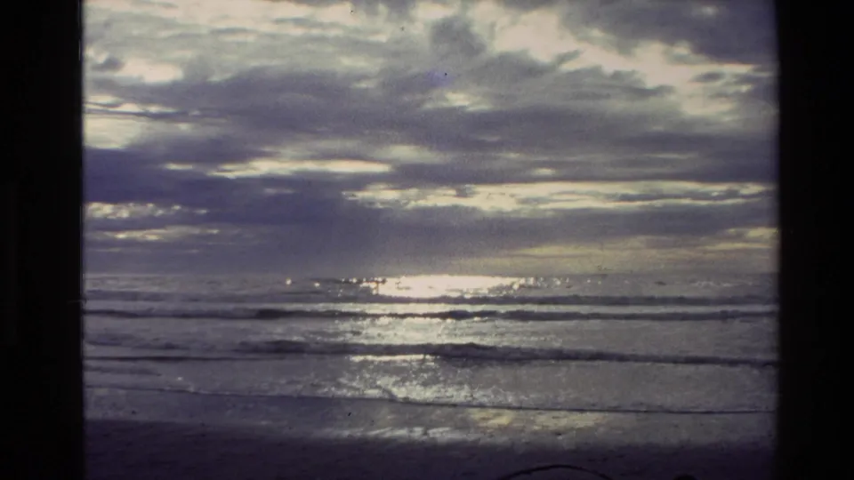 1982 New Zealand 8mm frame showing glints of sunlight on the water, layered clouds, and gentle surf near the shore.