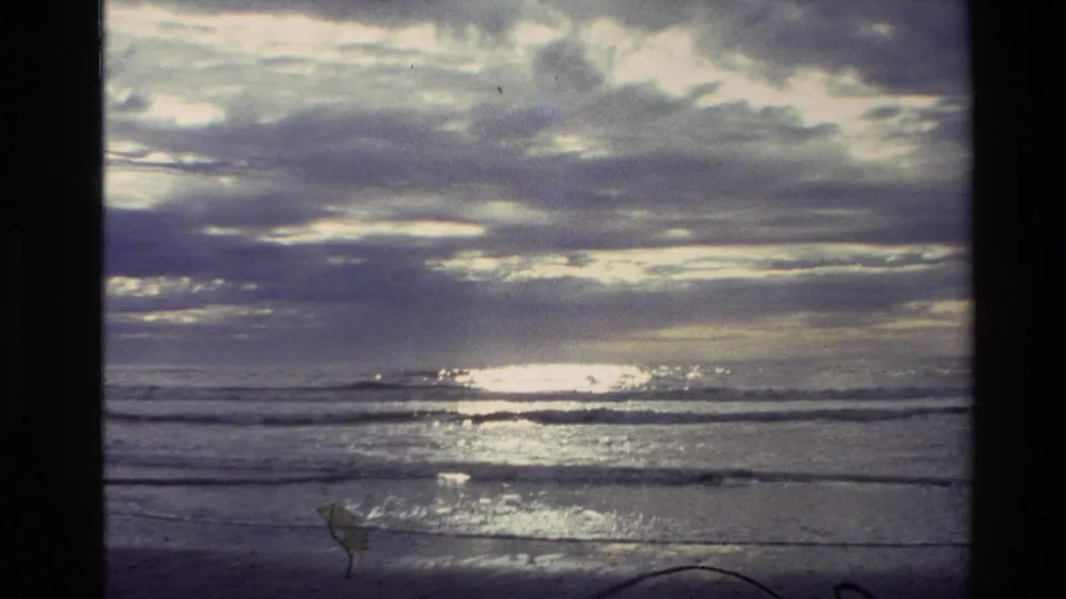 1982 New Zealand 8mm frame of a cloudy beach view with sunlight flashing on the sea and low waves crossing the foreground.