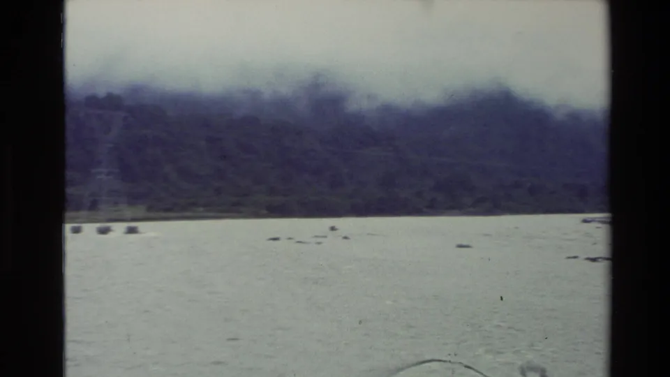 1982 New Zealand 8mm frame showing a wide riverbed beneath misty forested mountains and an overcast sky.