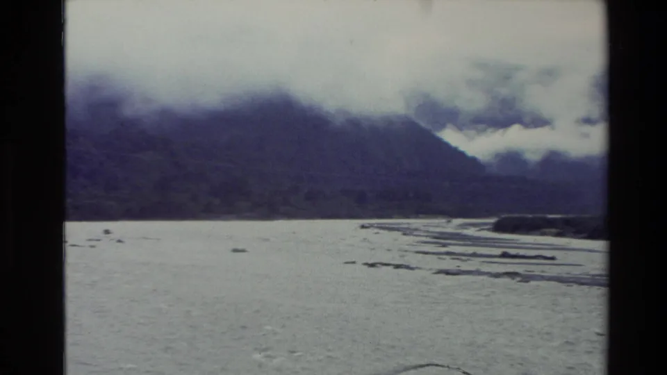New Zealand home-movie frame from 1982 with a broad river channel, exposed bars, and clouded mountain slopes.