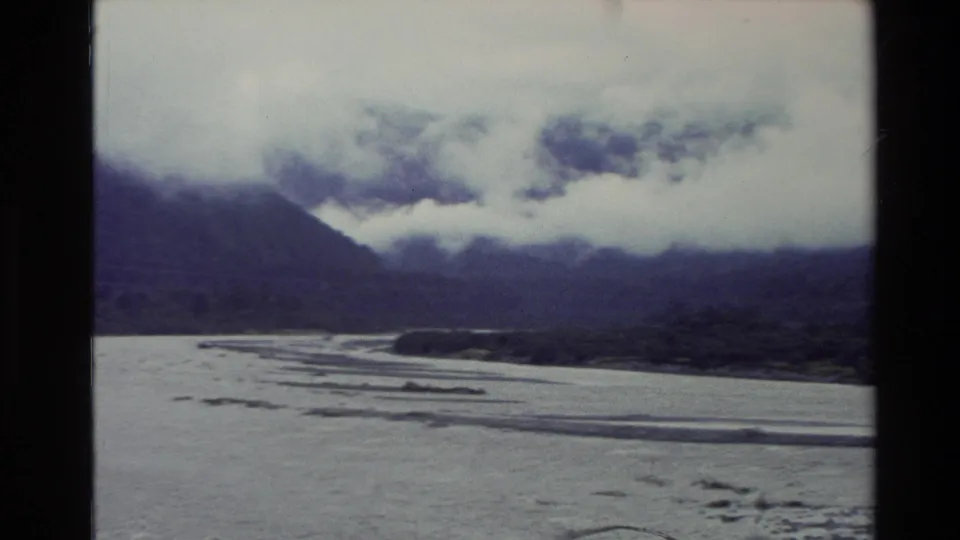 1982 New Zealand 8mm view of a river bend, pale sandbars, and a dark mountain ridge under low cloud.