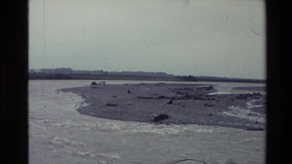 New Zealand 1982 home-movie frame showing a rocky riverbed, flowing water, gravel islands, and a low distant horizon.