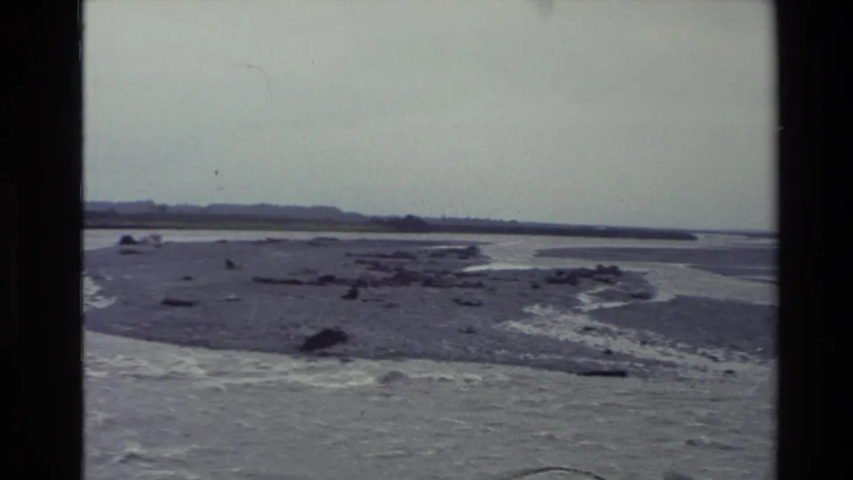 1982 New Zealand 8mm frame showing braided river flats, shallow water channels, and a hazy tree-lined horizon.