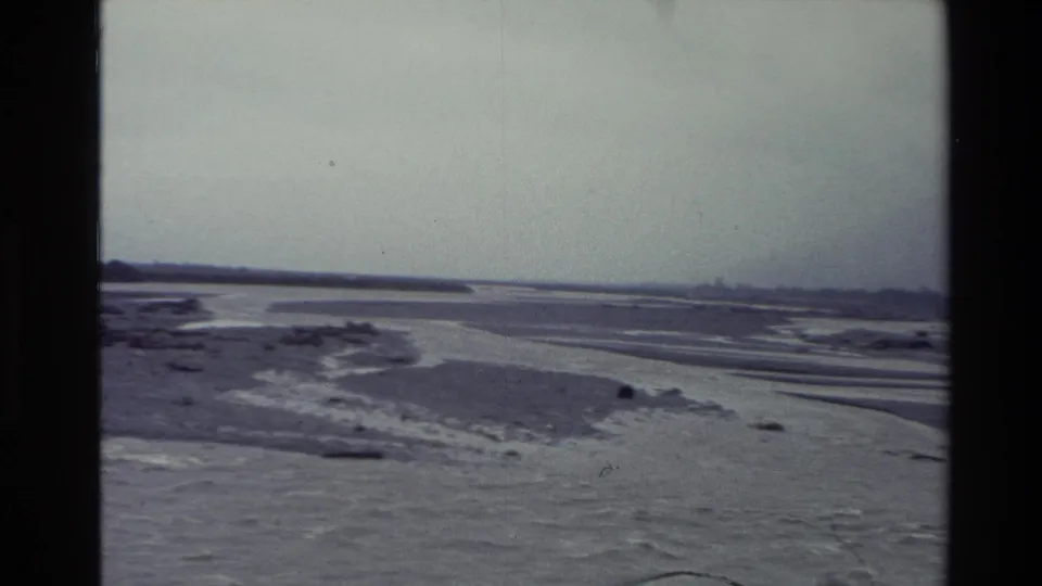 1982 New Zealand home-movie frame of a broad braided stream bed with winding water channels and a muted cloudy horizon.