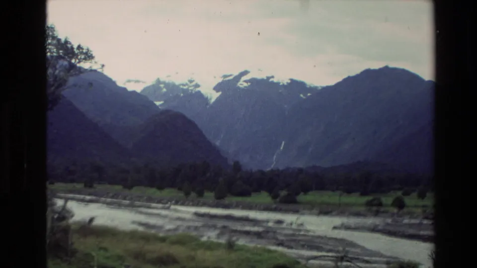 1982 New Zealand 8mm frame showing a stream in a mountain valley, dark slopes, green ground, and snow patches under cloud.