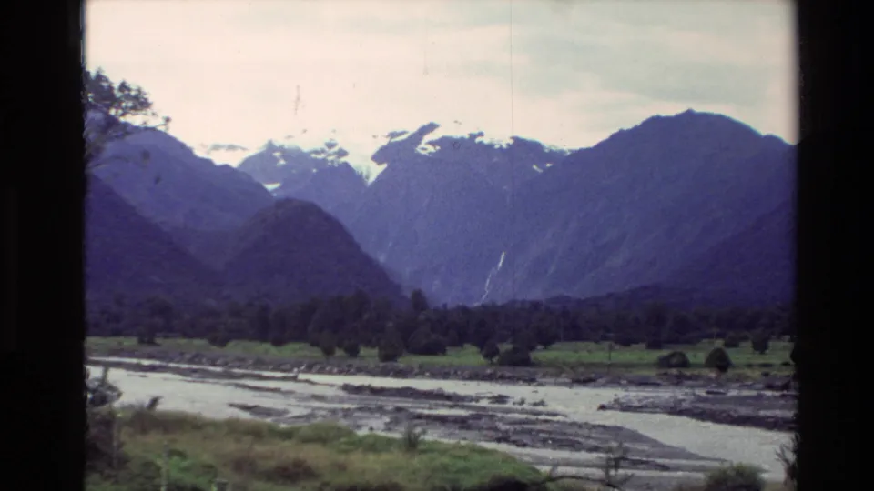 1982 New Zealand valley view with river channels, forest, green meadowland, and distant snowy peaks.