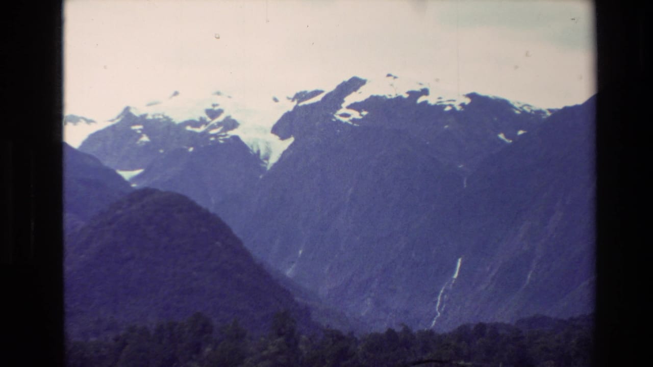 1982 New Zealand home-movie view of snow-capped alpine peaks beyond dark forested slopes, with soft 8mm color and frame edges.
