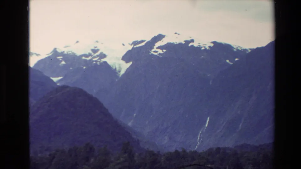 1982 New Zealand alpine landscape with snow patches on high peaks and a narrow waterfall descending a dark mountainside.