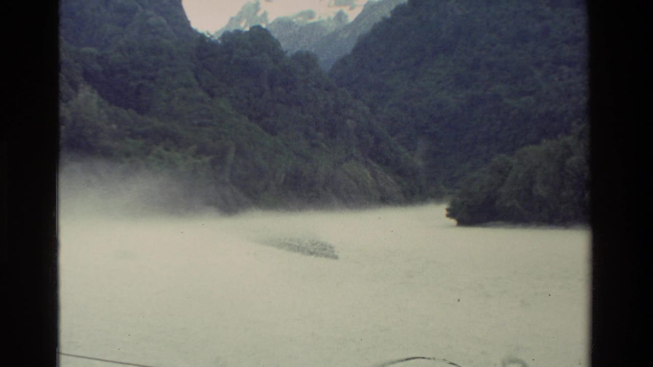Pale lake below dark forested mountains with mist along the shore.