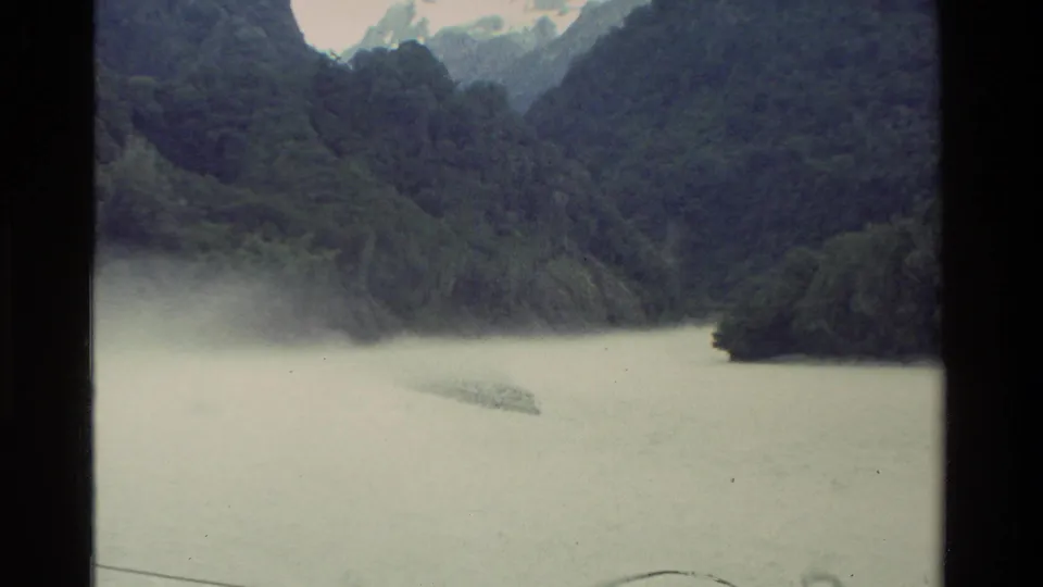 1982 New Zealand 8mm frame showing a calm lake bordered by steep forested mountains and distant snow patches.