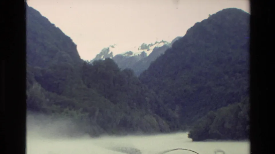 1982 New Zealand travel frame with dark green slopes, a calm lake edge, and snow-capped peaks beyond the valley.