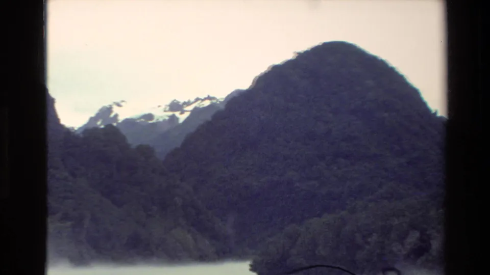 1982 New Zealand 8mm frame showing a rounded forested mountain, mist on the lake, and snow in the background.