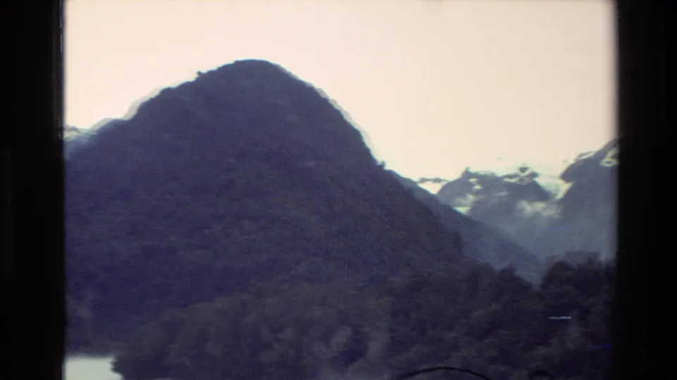 1982 New Zealand home-movie frame with a dark mountain foreground, snowy ridges behind it, and mist over the lake edge.