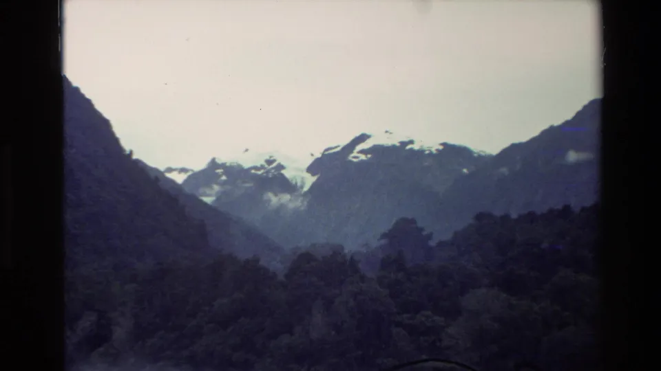 1982 New Zealand alpine view with snow-marked peaks, shadowed slopes, and dense forest in the lower frame.