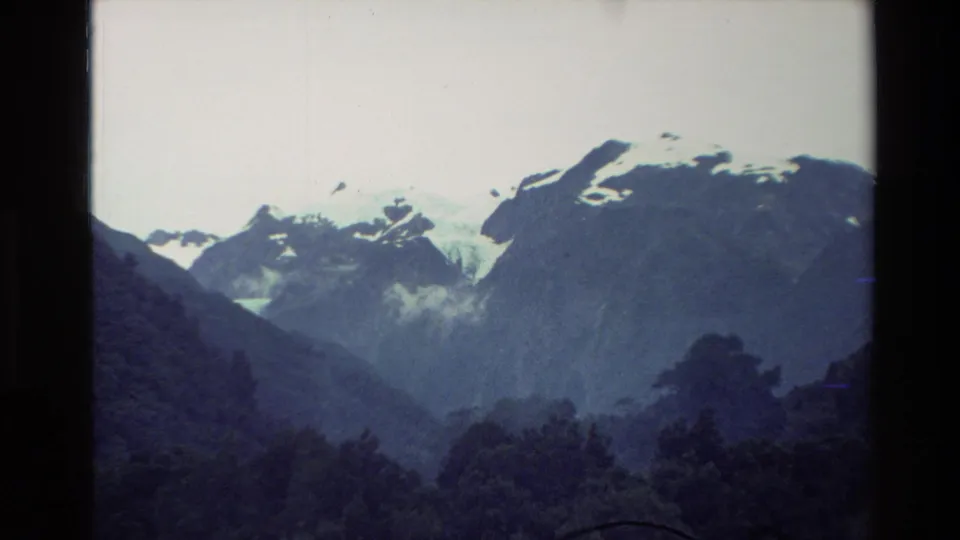 1982 New Zealand home-movie frame focusing on snow-marked mountain ridges above wooded foothills.