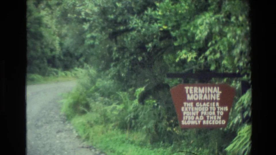 1982 New Zealand roadside frame showing a gravel road, dense vegetation, and a red Terminal Moraine glacier sign.