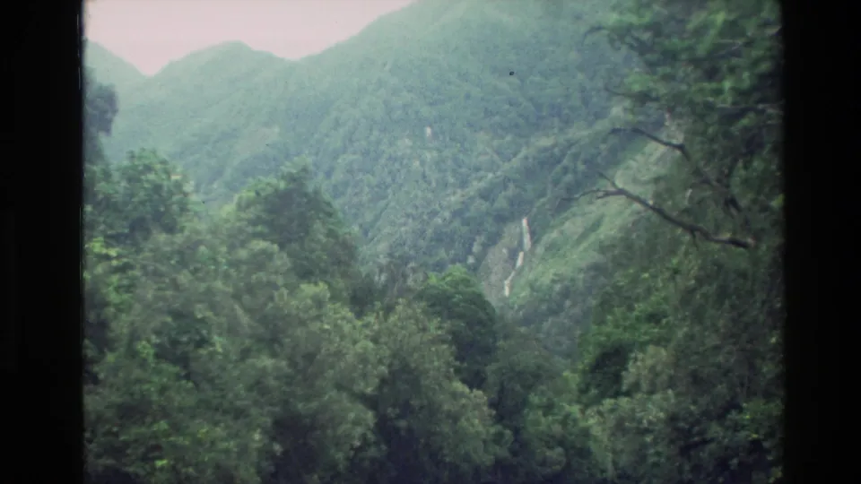 1982 New Zealand 8mm frame showing a green mountain valley, dense forest canopy, and a distant waterfall through dark frame edges.