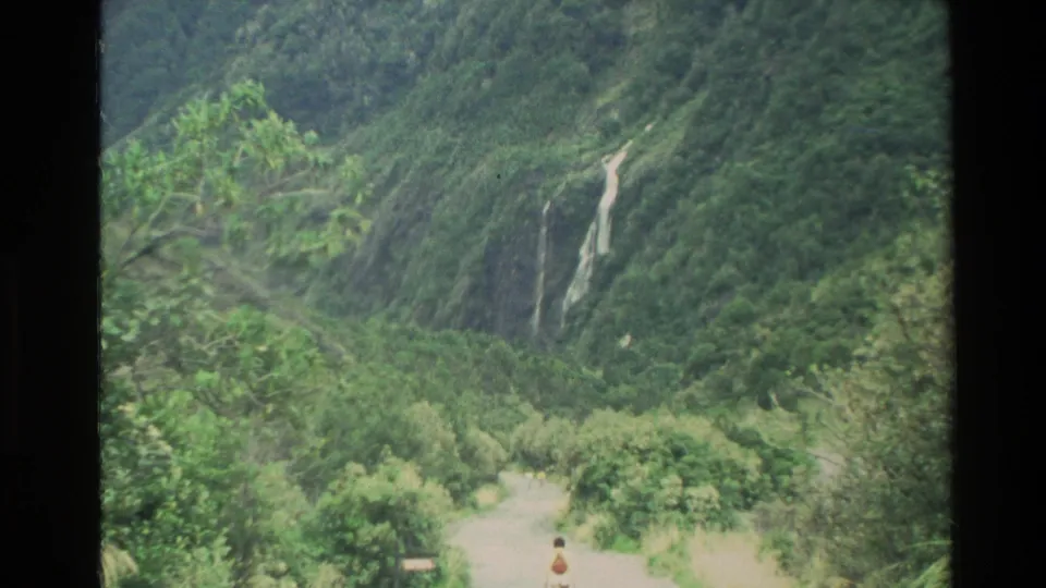 1982 New Zealand 8mm frame showing a distant person on a winding path below a waterfall and densely vegetated mountain slope.